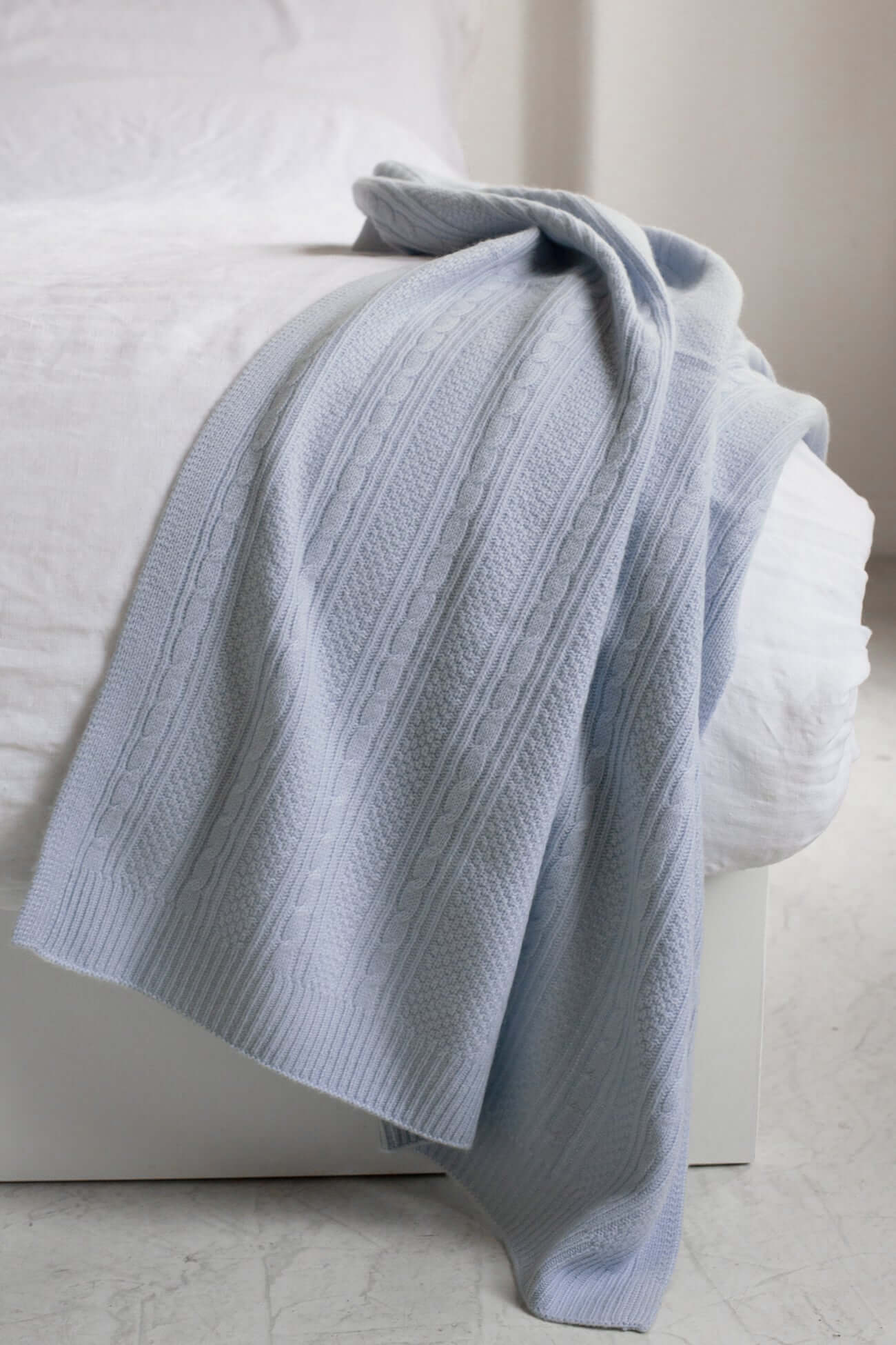 Soft blue knitted blanket draped over a white bed, adding a cozy touch to the accessories collection.